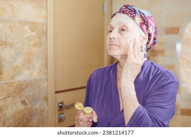 Elderly woman taking care of her face. Senior woman standing at the mirror in the bathroom and applying cream or mask on her face 库存照片