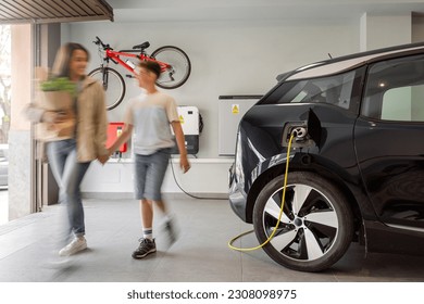 Electric vehicle charging station in private home with mother and son walking alongside, blurred family leaving the house, with a bicycle hanging on the wall. Horizontal., motion blur. 库存照片