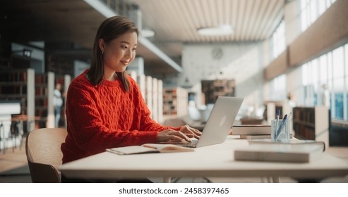Engaged Asian Female Student Working on Her University Assignments With a Laptop in Modern Library. Young Woman in Red Sweater Studying, Surrounded by Books and Pens. Adlı Stok Fotoğraf