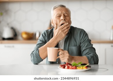 日中の過度の眠気。疲れた年配の男性が台所のテーブルであくびをし、年配の紳士が家で昼食を食べながら眠気を感じ、手で口を覆い、過眠症を患っているの写真素材