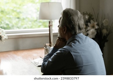Far away in thoughts. Rear view of aged hoary male sit by table at home alone look at window ponder prop chin on folded hands. Thoughtful mature man going down to memories reviewing past. Copy space Stock fotografie