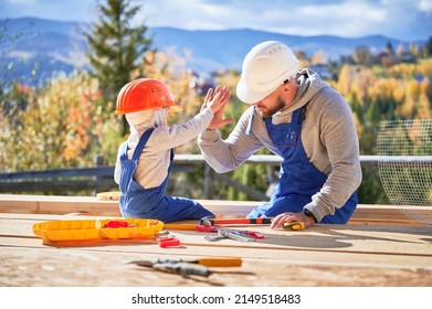 Father with toddler son building wooden frame house. Male builder giving high five to kid on construction site, wearing helmet and blue overalls on sunny day. Carpentry and family concept. Stock Photo