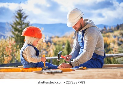 Father with toddler son building wooden frame house. Male builders hammering nail into plank on construction site, wearing helmet and blue overalls on sunny day. Carpentry and family concept. Stock Photo