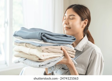 부드러움과 위생을 느껴보세요. 스마일 아시아 젊은 여자 터치 신선한 깨끗한 더미를 냄새 푹신한 흰색 셔츠, 스택 옷, 빨래 후 편안한 냄새. 집안일, 하녀 개념의 집안일. 스톡 사진