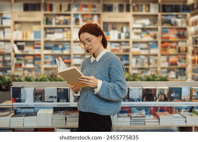 A focused young woman in glasses and a blue sweater reads a book while standing in a vibrant, well-stocked bookstore. The shelves are filled with a variety of books. स्टॉक फ़ोटो