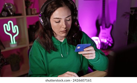 A focused young woman with headphones in a neon-lit gaming room examines a credit card at night. ภาพถ่ายสต็อก