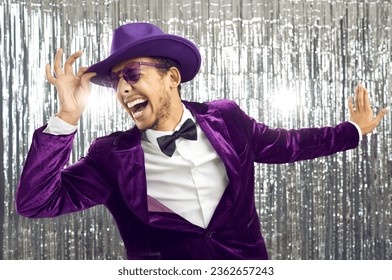 Funny happy excited crazy confident handsome man wearing purple velvet suit, bow tie, glasses and fedora hat having fun, singing loud songs and dancing against shiny foil fringe background at party 库存照片