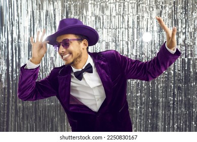 Funny smiling eccentric guy having fun at a party. Happy goofy confident man wearing a purple velvet suit, glasses, bow tie and fedora hat dancing against a shiny silver foil fringe background 库存照片