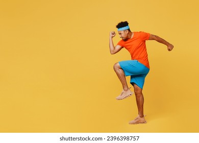 Full body side view young fitness trainer sporty man sportsman wear orange t-shirt raise up leg, ready to run spend time in home gym isolated on plain yellow background. Workout sport fit abs concept Foto Stock
