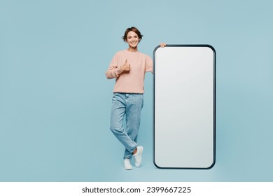 Стоковая фотография: Full body young fun woman she wear beige knitted sweater casual clothes big huge blank screen mobile cell phone smartphone with workspace area show thumb up isolated on plain pastel blue background