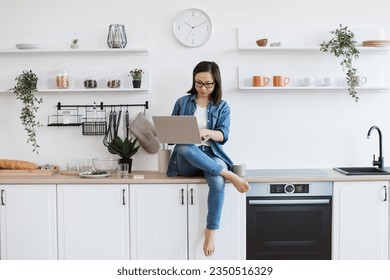 Full length view of asian woman with computer on knee sitting comfortably on worktop in stylish kitchen. Focused young student in glasses reading online article about new career options at home. 库存照片