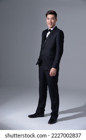 Full length portrait of a young businessman in wear formal black suit with bow tie on gray background 库存照片
