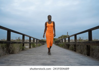 Full length of positive black lady in orange button down gown walking on footbridge and looking away against mountains and green bushes on background, fotografie de stoc