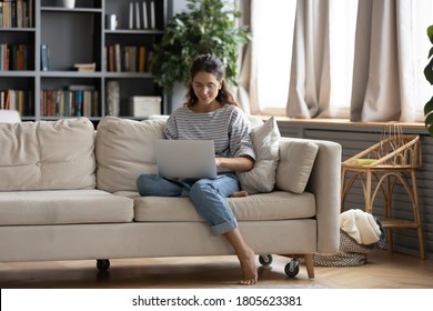 眼鏡をかけた若い魅力的な裸足の若い女性が、ソファーに座り、パソコンを使ってソファーに座り、自宅から離れて仕事を楽しみ、ノートパソコンのアプリを使ったり、オンラインで買い物をしたり、ネットサーフィンを楽しんだりする。の写真素材
