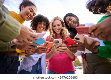 Group of international happy generation z only women laughing gathered in circle looking at cell phones outdoors. Cheerful young females using mobiles with funny expression. Addiction to technology  Foto Stock