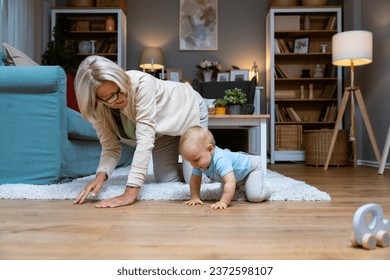 Grandmother of single mother taking care of her young baby, playing on the apartment floor. Senior woman babysitting her grandson or granddaughter while child parent are at work or date night. 库存照片