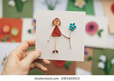 Hand holding quilling card with stick figure girl giving flower. woman making greeting cards. Hand made of paper quilling technique. Handicraft at home. Hobby, home office. Stock-foto