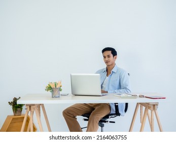 Handsome young Asian man in denim shirt using laptop on table on white clean space wall background in home office. Learning, working, startup with small business, stock, financial, investment concept. Adlı Stok Fotoğraf
