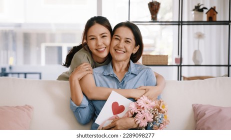 Happy time Mother day grown up child looking at camera cuddle hug give flower gift box red heart card to mature mum. Love kiss care mom asia middle age adult people smile enjoy sitting at home sofa. Stock Photo