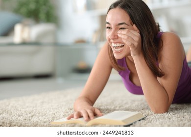 家の床に横たわっているコメディーの紙の本を陽気に読んで笑っている幸せな女性の写真素材