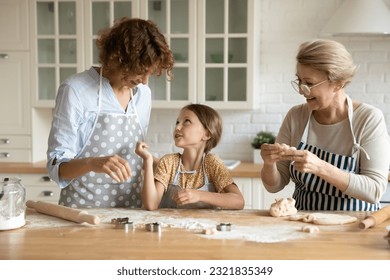 幸せな若い35sの女性は楽しい小さな就学前の子供の娘と一緒に手作りのケーキを作り、現代のキッチンで美しい高齢の退職母を気遣って楽しく話しています，家族の料理活動。の写真素材