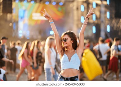 Happy young woman enjoys summer music festival on beach. Arms raised, peace sign, dancing in crowd with stage lights. Casual fashion, sunglasses, fun with friends at sunset concert party. 库存照片
