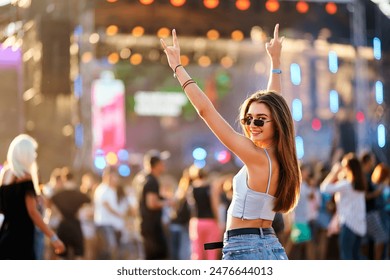 Happy young woman enjoys summer music fest, raises hands with peace sign, dances at beach party. Vibrant festival atmosphere, dusk light with stage lights. Fashionable crowd, holiday vibes, fun. 库存照片