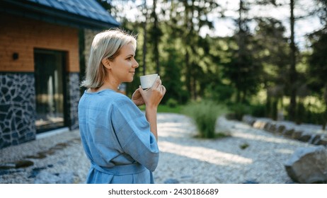 コテージの近くの庭に立って、山での夏休みに朝のコーヒーを楽しんでいる幸せな若い女性。の写真素材