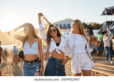 Happy young women dance at beach music fest, sunset creates perfect party atmosphere. Friends enjoy live bands, summer vibes by sea. Casual festival fashion trends, unforgettable moments together. 库存照片