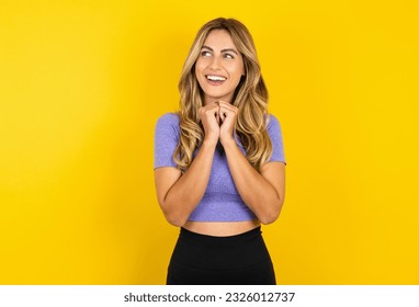 Heureuse Jeune belle femme blonde portant un vêtements de sport sur fond jaune studio anticipe quelque chose de génial, semble joyeusement à l'écart, garde les mains ensemble près du visage, a une expression heureuse. : photo de stock