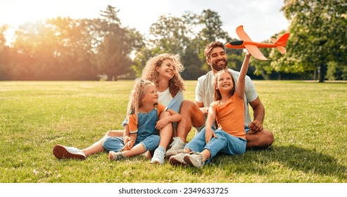 행복한 어린 가족 아빠, 엄마 그리고 두 명의 딸들이 따뜻한 화창한 날에 공원의 풀밭에 있는 초원에 앉아 하루를 쉬면서 즐거운 시간을 보내며 비행기를 가지고 놀고 있습니다. 스톡 사진