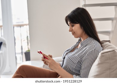 Happy young indian woman using smart phone sitting on couch at home. Smiling lady holding cellphone doing online shopping, ordering delivery in smartphone application, subscribing new social media. 库存照片
