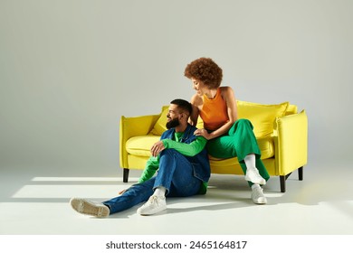 A happy African American man and woman in vibrant clothes sit together on a yellow couch against a grey background. ภาพถ่ายสต็อก