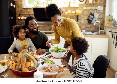 Happy African American woman having Thanksgiving lunch with her family and serving salad at dining table.  Stock Photo