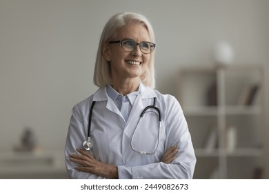Happy attractive older attending physician in white coat posing in clinic office, smile, look into distance, feel satisfied with workday and professional aid to patients. Mission, profession, medicine Stock-foto