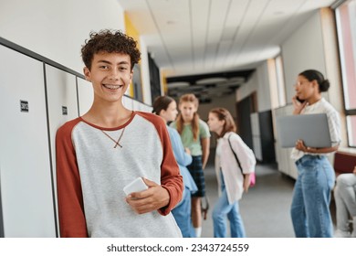 happy boy with braces holding smartphone and looking at camera during break in school hallway Foto stock