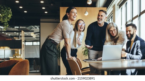 Happy businesspeople laughing while collaborating on a new project in an office. Group of diverse businesspeople using a laptop while working together in a modern workspace. ஸ்டாக் ஃபோட்டோ