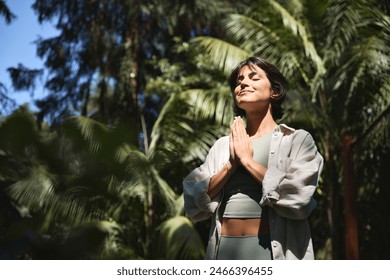 Happy calm young Hispanic woman holding hands in namaste meditating doing yoga breathing exercises with eyes closed feeling peace of mind, mental balance standing in green nature tropical park. 库存照片