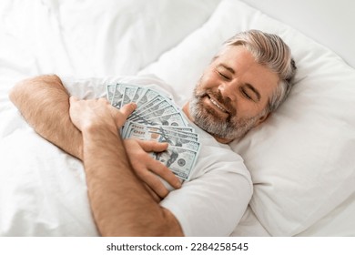 Happy cheerful handsome grey-haired bearded middle aged man wearing pajamas lying in bed with closed eyes, embracing cash dollar banknotes and smiling, top view. Money savings, financial literacy 库存照片