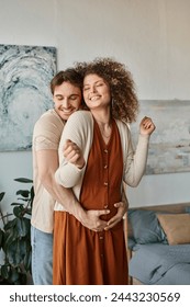 赤ちゃんを待つ幸せなカップル、男は陽気な笑顔で後ろから彼の妊娠中の妻を抱きしめていますの写真素材