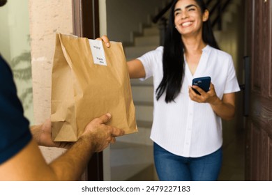Happy latin woman receiving a food delivery order in a brown paper bag at the doorway of her home 库存照片