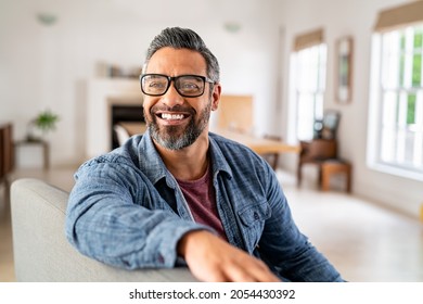 Happy mature middle eastern man wearing eyeglasses sitting on couch. Portrait of indian man relaxing at home and looking away with big smile. Mid adult guy with specs thinking about his future., fotografie de stoc