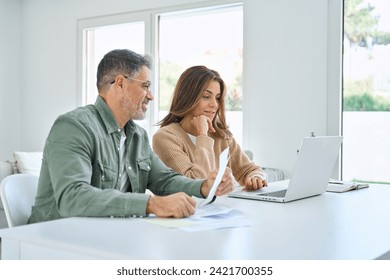 Happy middle aged mature man and woman paying bills online at home. Older senior couple using laptop computer checking insurance or financial invoice counting taxes sitting at table in living room. 庫存照片