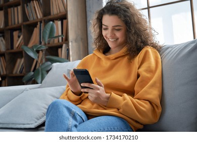 快乐的千禧一代西班牙裔青少年女孩在家里拿着智能手机检查社交媒体.微笑的年轻拉丁女人使用手机应用程序玩游戏，在线购物，订购送货在沙发上放松。 库存照片