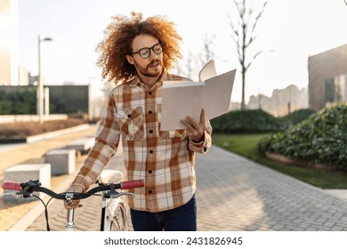 Happy redhead man reading book near bicycle on footpath स्टॉक फ़ोटो