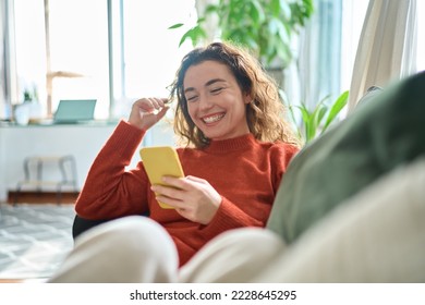 携帯電話を使ってソファに座っている幸せなリラックスした若い女性、スマートフォンを持って笑っている笑顔の女性、携帯電話を見て、モバイルアプリでオンラインEコマースショッピングをしたり、ビデオを見たりして楽しんでいる。の写真素材
