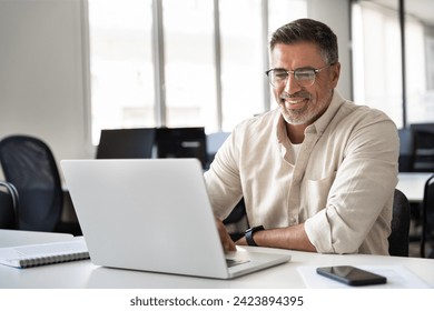 Happy smiling mature Indian or Latin business man ceo trader using computer, typing, working in modern office, doing online data market analysis, thinking planning tech strategy looking at laptop. Adlı Stok Fotoğraf