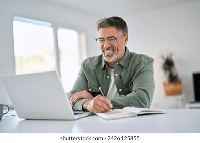 Happy smiling middle aged older mature man wearing glasses looking at laptop using computer writing notes watching webinar sitting at home table. Hybrid work, elearning, online learning concept. Adlı Stok Fotoğraf