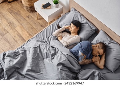 ベッドの上に横たわっているアジアの妻と入れ墨の男の高角度ビュー、関係の困難の概念の写真素材