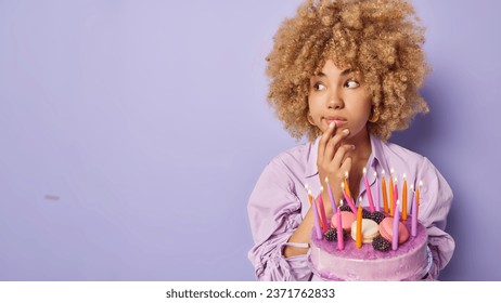 Horizontal shot of beautiful curly haired woman keeps hand on chin concentrated aside poses with tasty cake and burning candles celebrates special occasion isolated over purple background copy space 库存照片